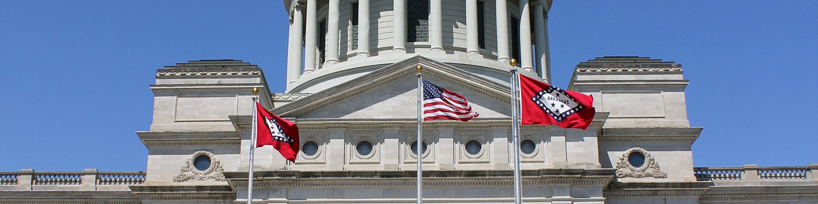 The Arkansas state flag (x2) and the U.S. flag flying in front of the Arkansas Capitol Building.