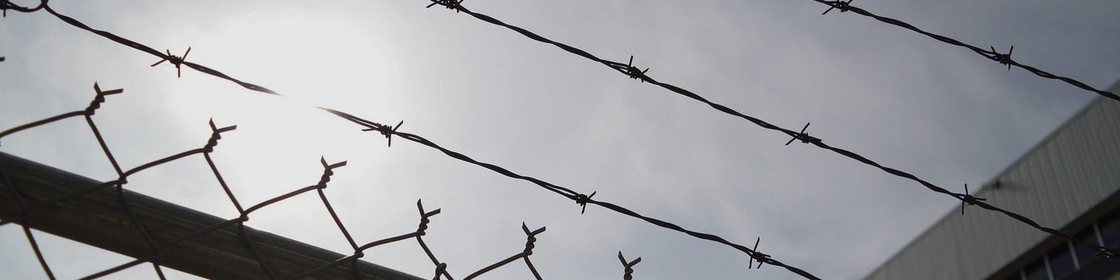 The sun peeking through clouds behind a chainlink/barbed wire fence.